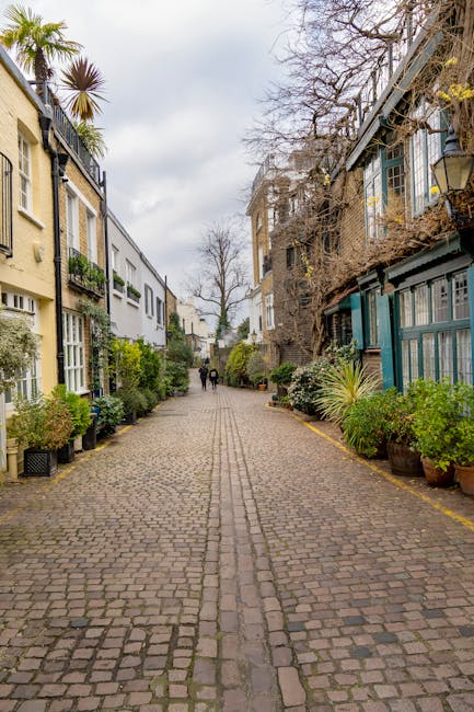 A cobblestone street in West Kensington North End Road, lined on both sides by residential buildings with garden areas. On the left, a yellow and white house features potted plants and greenery along its front, while on the right, a building with teal window frames and a garden with various plants and shrubs. Sparse trees with bare branches are visible overhead, indicating a cooler season, and the street is partly wet, suggesting recent rain. In the background, a few pedestrians are walking along the pathway, with some maintaining the surrounding greenery, indicative of garden maintenance work. The overall scene emphasizes well-kept outdoor spaces and thoughtful landscaping, with a focus on garden and outdoor area upkeep by Gardeners West Kensington.