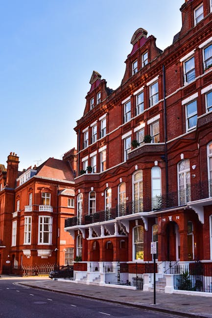 The image shows a historic red brick building with ornate architectural details, including decorative cornices and arched windows, situated on a tree-lined street. In front of the building, there is a small front garden area with neatly maintained grass, a gravel pathway, and a low black metal fence. The garden features a variety of plants, including small shrubs and flowering perennials, which appear well-kept. Next to the garden, a paved footpath runs alongside the street, with a parking sign and a few parked cars visible. The photograph is taken during daylight under clear blue skies, highlighting the vibrant color of the brickwork and the lush greenery, suggesting a well-maintained outdoor space managed by professional gardening services such as Gardeners West Kensington, who specialize in garden maintenance and landscaping work for homes on North End Road. The scene emphasizes the tidy, landscaped frontage that complements the elegant exterior of the building.