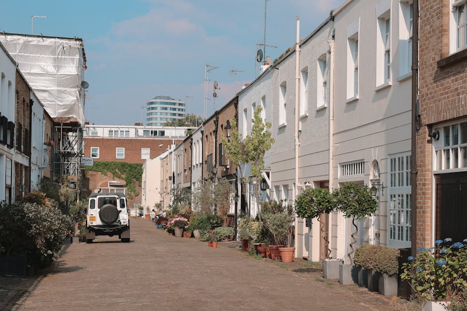 A cobblestone street in West Kensington North End Road, lined on both sides by residential buildings with garden areas. On the left, a yellow and white house features potted plants and greenery along its front, while on the right, a building with teal window frames and a garden with various plants and shrubs. Sparse trees with bare branches are visible overhead, indicating a cooler season, and the street is partly wet, suggesting recent rain. In the background, a few pedestrians are walking along the pathway, with some maintaining the surrounding greenery, indicative of garden maintenance work. The overall scene emphasizes well-kept outdoor spaces and thoughtful landscaping, with a focus on garden and outdoor area upkeep by Gardeners West Kensington.